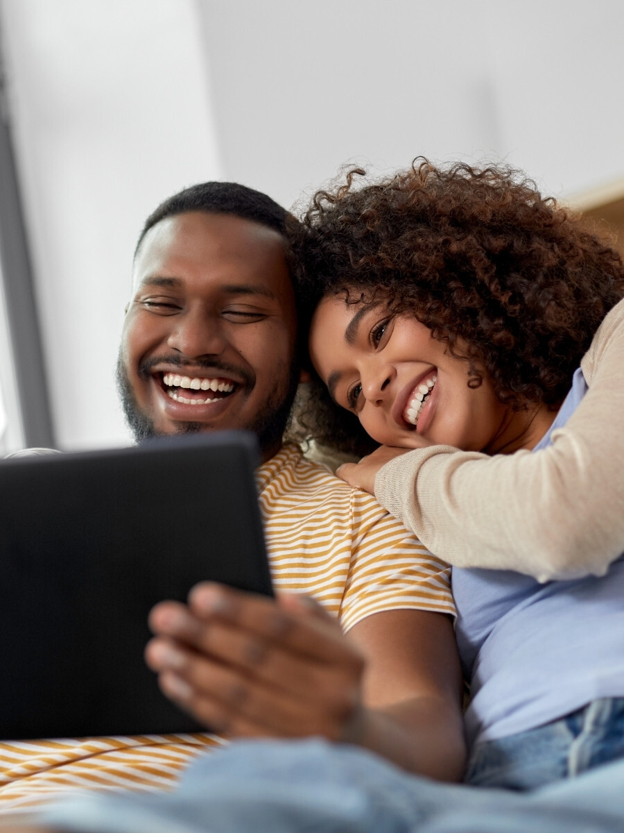 Couple sitting together on a couch, smiling and looking at a tablet for a virtual counseling session in a relaxed, supportive home environment associated with couples counseling.