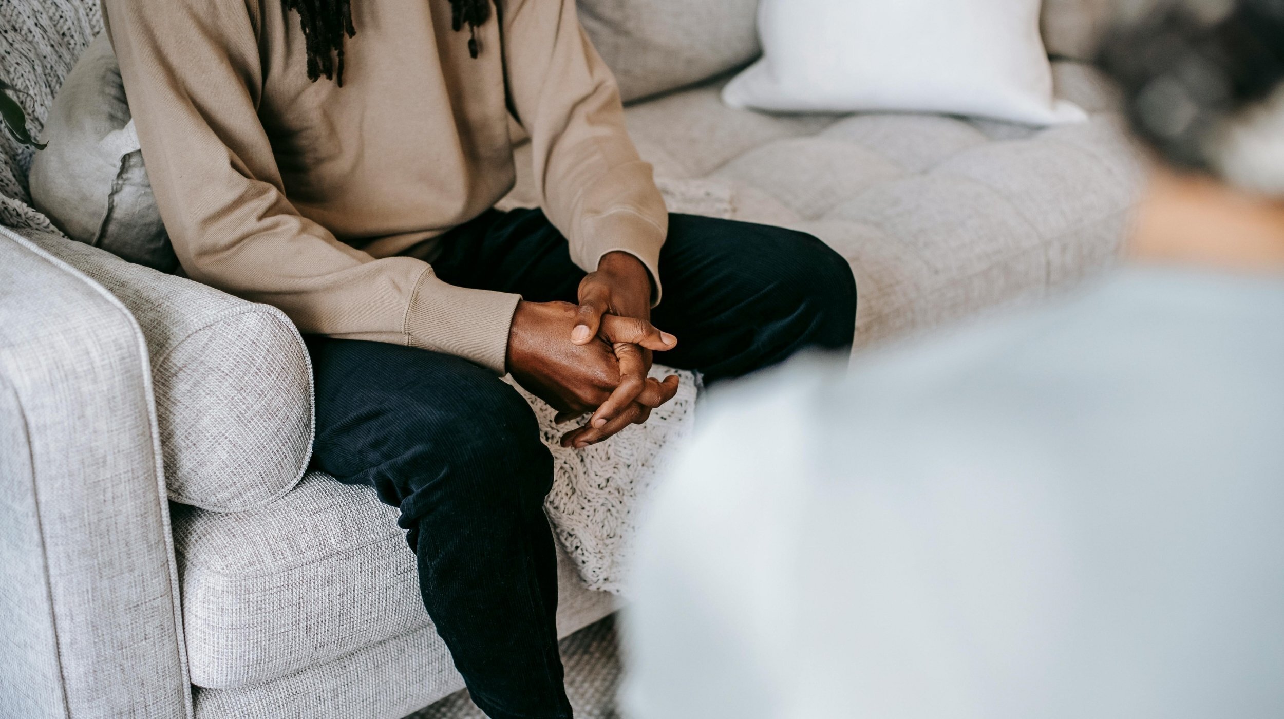 Person seated on a couch with hands clasped, appearing thoughtful during a counseling session in a calm indoor setting.